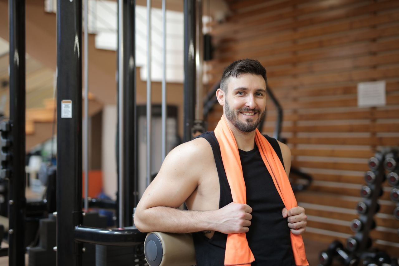 Smiling man poses in gym, showcasing fitness and strength with gym equipment.